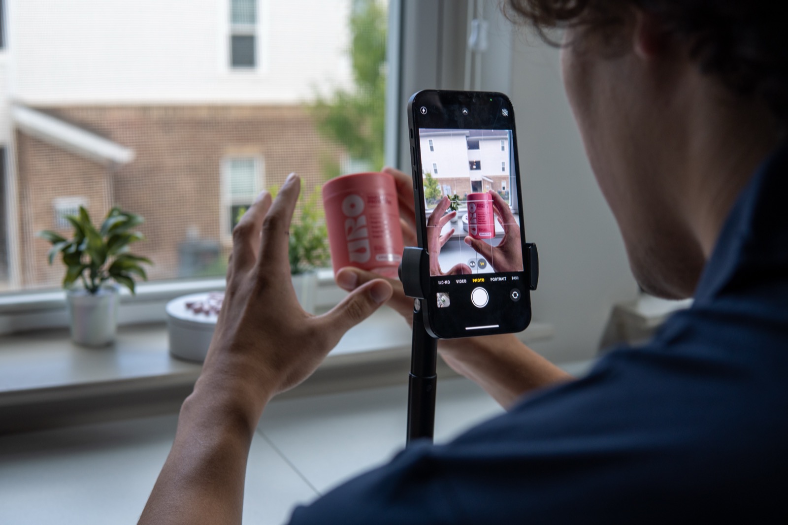 Operator framing a product on a tripod-mounted phone for a TikTok live broadcast, daylight from a tall window behind.